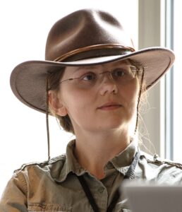Becky Yoose with her signature brown cowboy hat in front of a sun-lit glass window, wearing thin-rimmed glasses and green-grey safari shirt. Her face is angled toward the right. 
