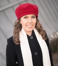 Heidi Blackburn wears black winter coat, red beret, and a long white scarf loosely draped on her neck. She has long, brown wavy hair and smiles at the camera.