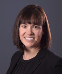 Megan Oakleaf with chin-length dark brown hair and sporting a straight bang, wearing a black suit. Her body is angled slightly to the left, smiling at the camera.