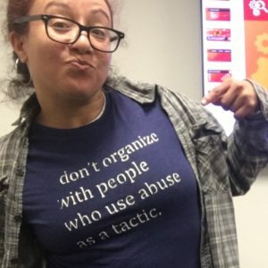 Myna Morales has brown curly hair and wears dark-rimmed square glasses. She’s doing a playful pose with her index finger pointed to her dark blue shirt with text “don’t organize with people who use abuse as a tactic”. She also wears a brown-green plaid shirt as a top layer.
