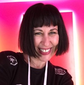 Paige Dansinger with dark chin-length, bob-cut hair style and wearing a dark shirt with some patterns in front of a neon pink, orange, and white background. Paige's body is slightly tilted to the right with a happy smile.