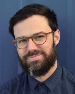 Scot Young with short, dark hair, thick mustache and full beard with some gray hair streaks on the chin, wearing tortoise-rimmed glasses. His face is tilted slightly to the left with a subtle smile toward the camera.
