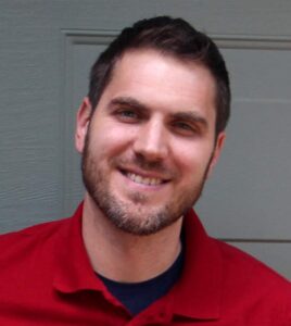 Sean Aery with very short, dark hair, mustache, and light beard around his chin and side face. He wears a deep red shirt and smiles at the camera with a head slightly tilted to the right.